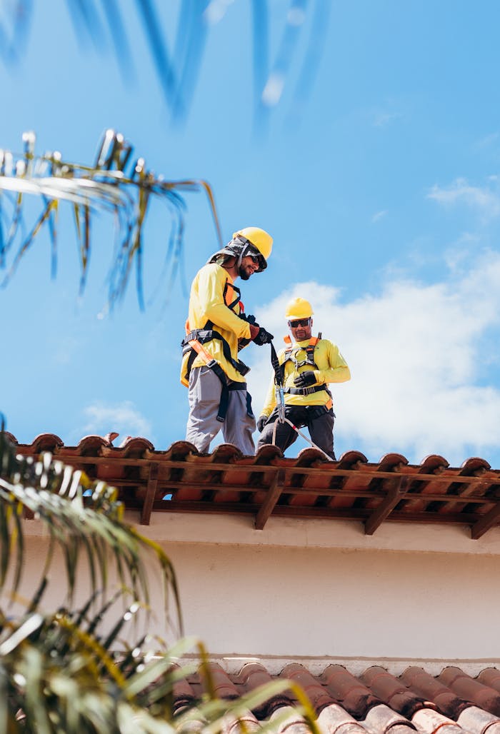 Two construction workers in safety gear working on a tiled roof under a clear blue sky.